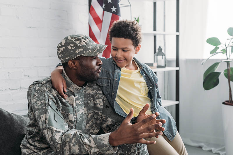 A man in military uniform sits on a couch with a child who has their arm around his shoulders. They are smiling and talking, with an American flag in the background.
