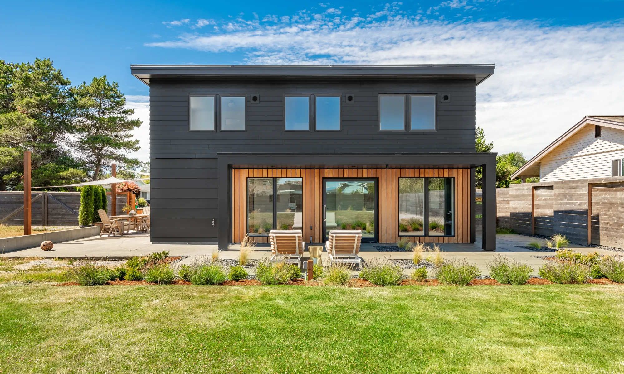 Modern two-story house with dark siding and large windows, wooden accents around the doors, a patio with two lounge chairs, and a landscaped yard with green grass and garden beds under a blue sky.