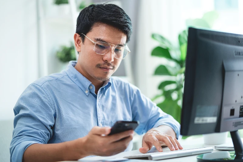 A man wearing glasses is sitting at a desk with computer monitor and cell phone